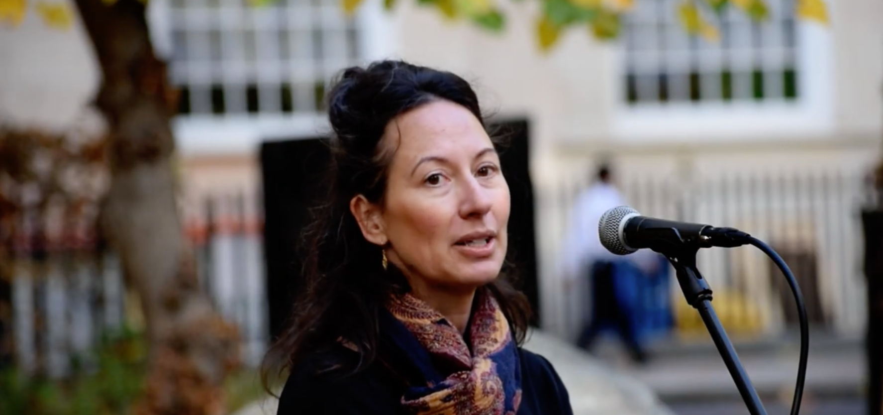 A woman with dark hair, wearing a scarf, speaking in to a microphone.
